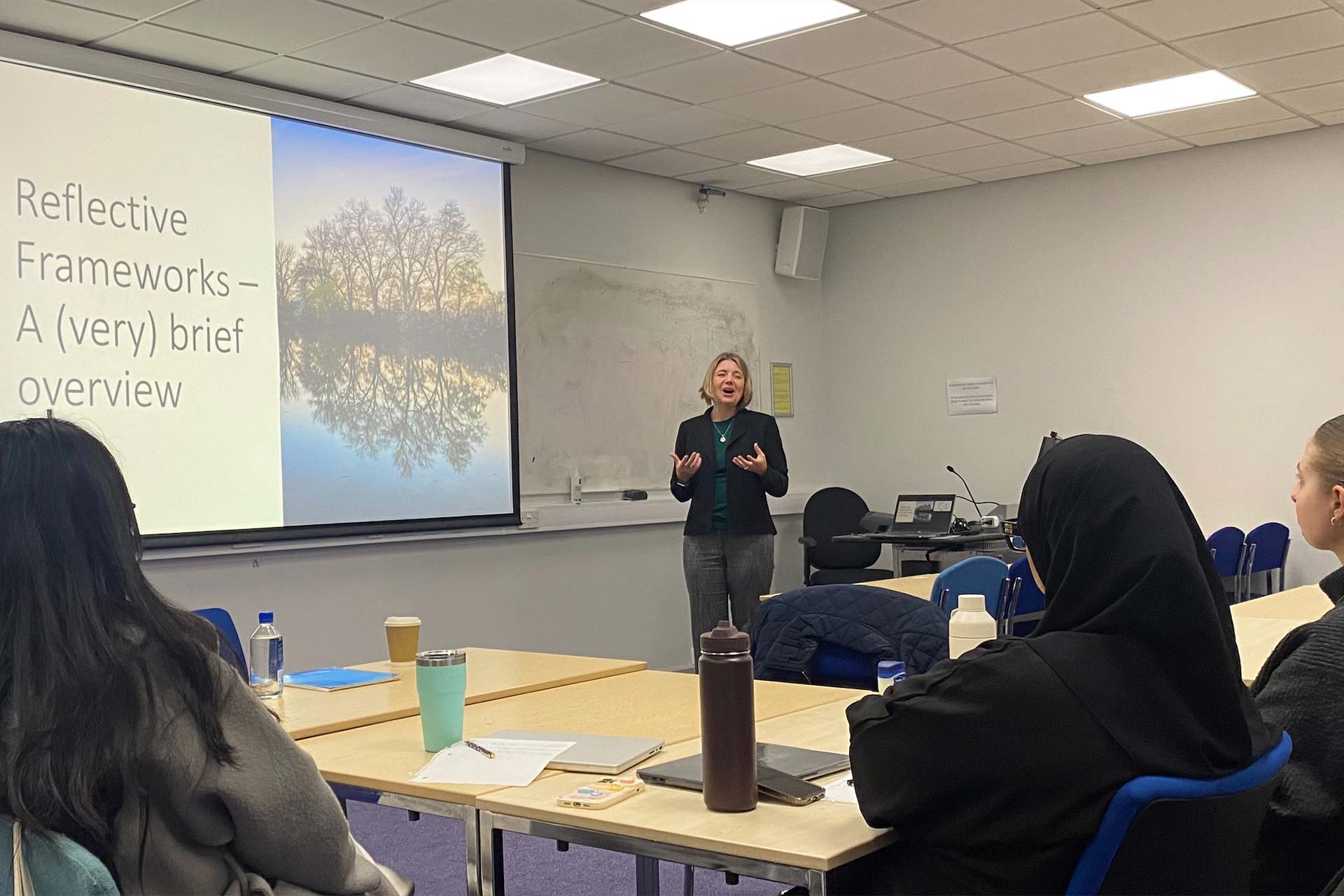 A woman with short blonde hair standing at the front of a classroom presenting to a group of people seated, the projected screen reads 'Reflective frameworks - a very brief overview