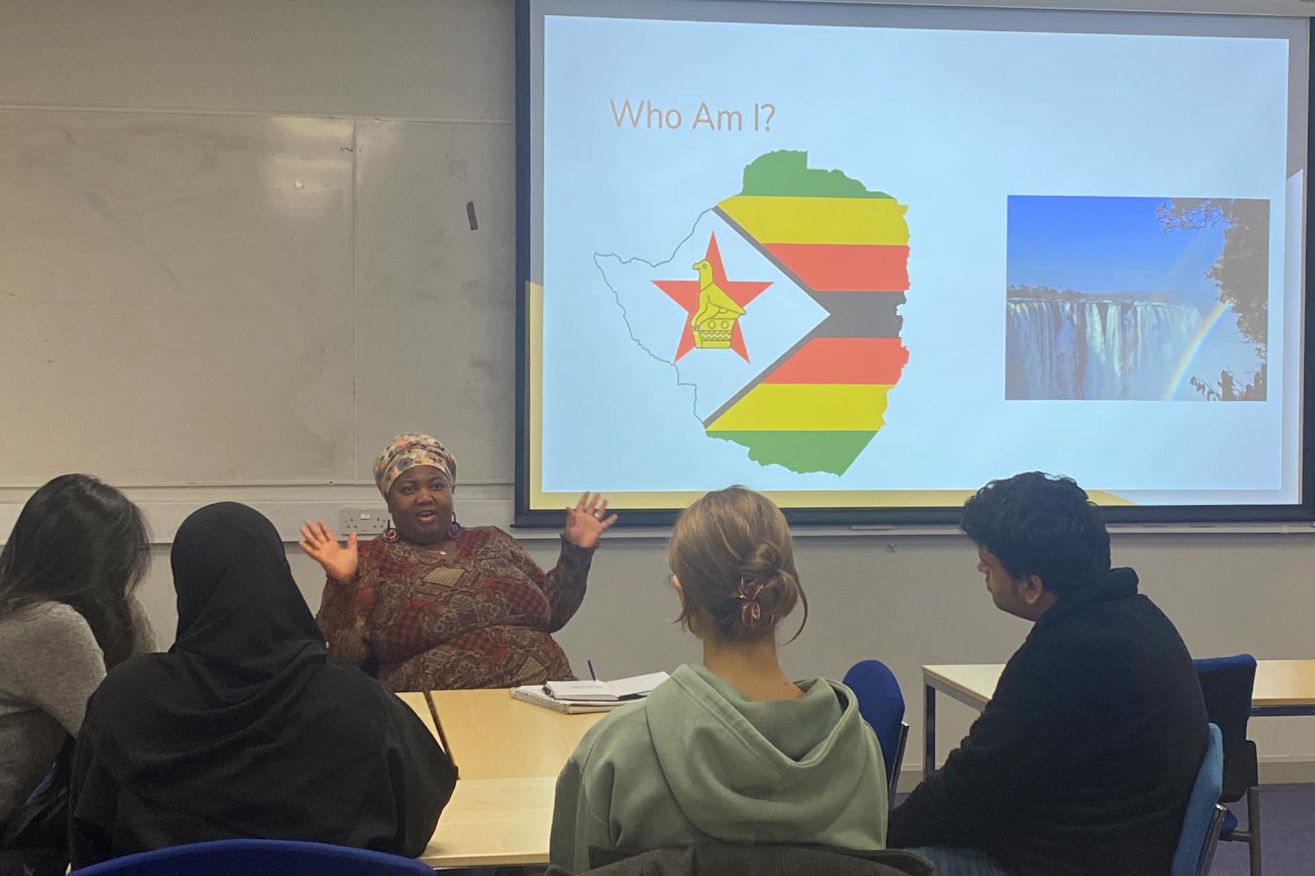 Tracy Mpofu, a Black woman, sits at a table presenting in front of a slideshow that reads 'Who Am I?' with a picture of a flag on it. Four students sit across the table listening to her opposite.