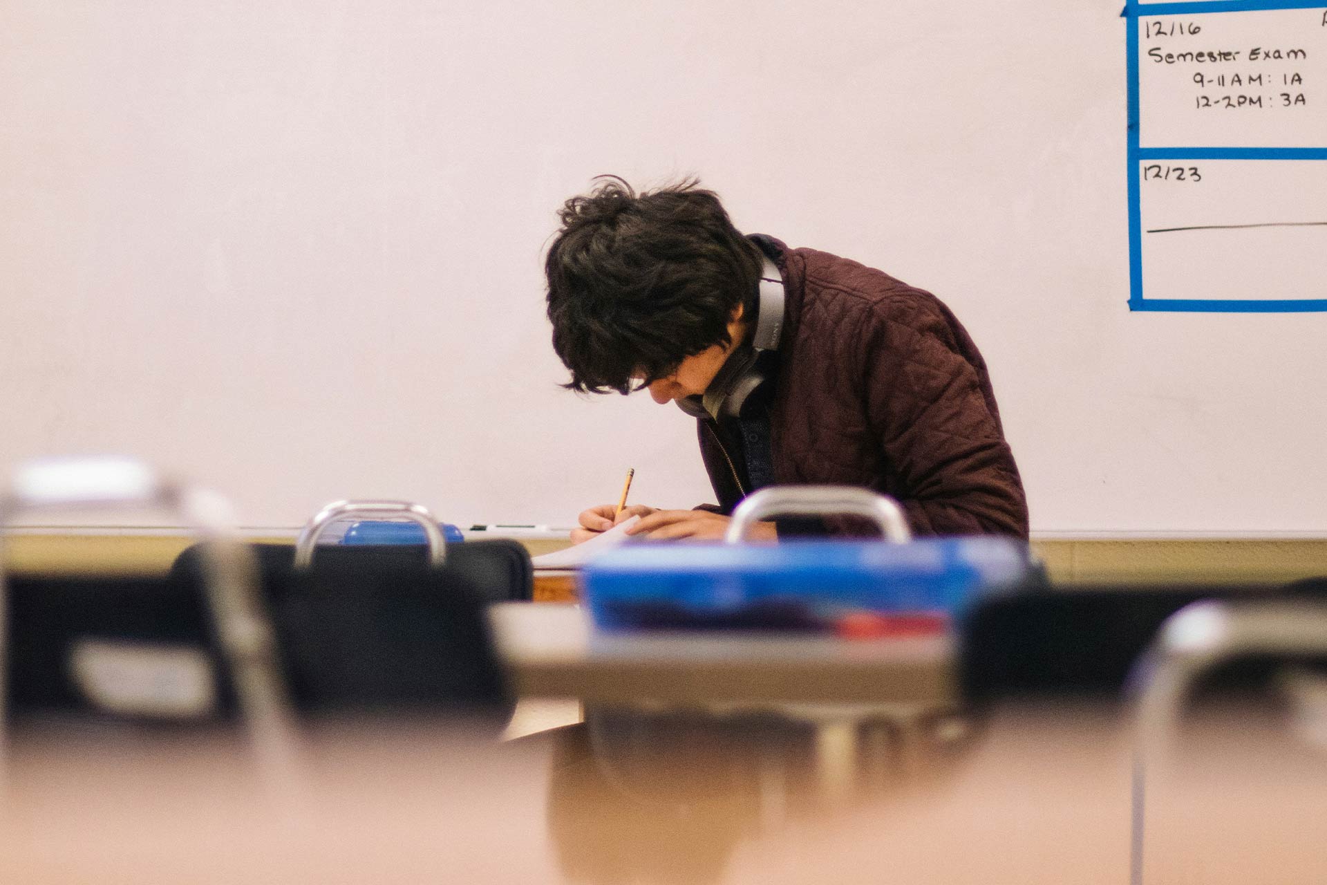 A young man sitting at a desk in a classroom writing on a piece of paper.