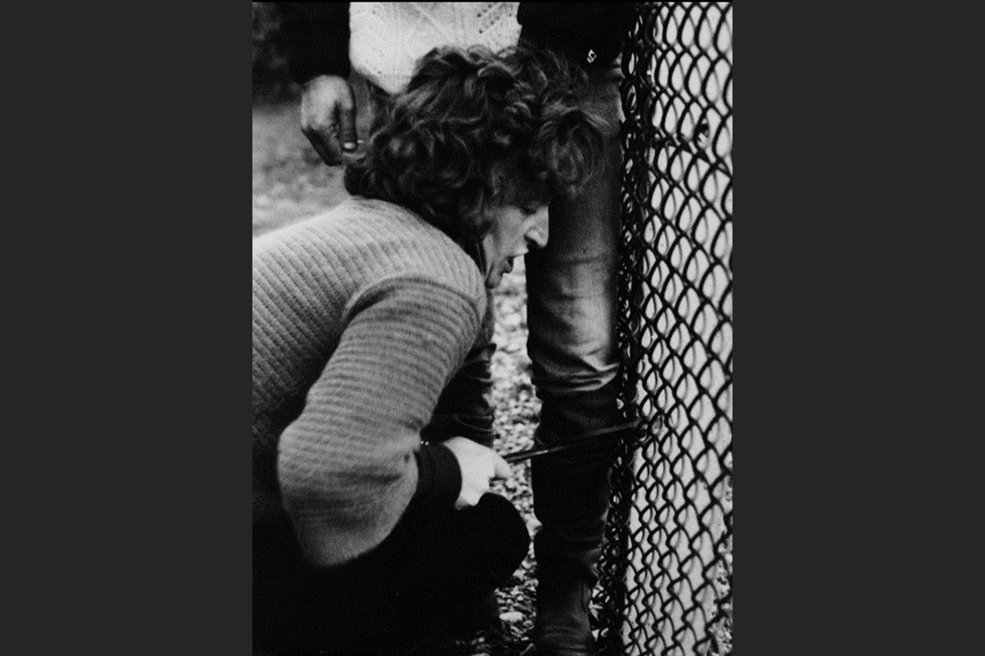 A black and white image of a woman crouching near a barbed wire fence with a sharp object in her hand.