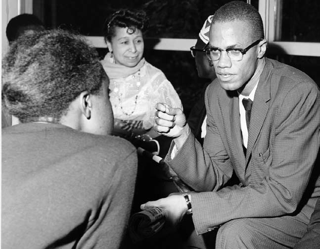 Three Black men sitting down in discussion, on the far right sits Malcolm X, wearing a suit and glasses, using his hand to gesticulate.