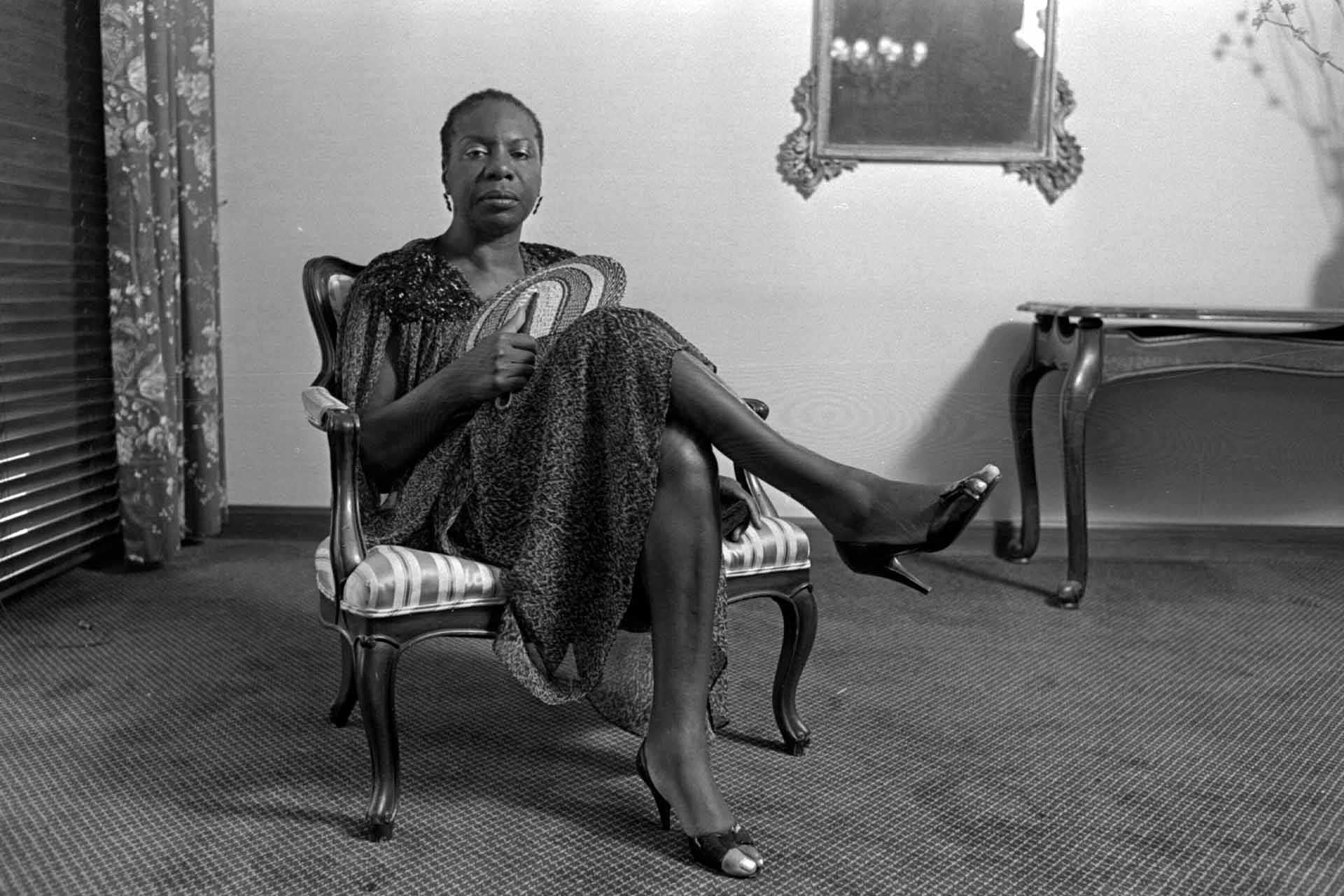 Nina Simone is a Black woman, she sits on a chair in a room inside a house or apartment, with her legs crossed and a fan in her hand in a black and white photograph.