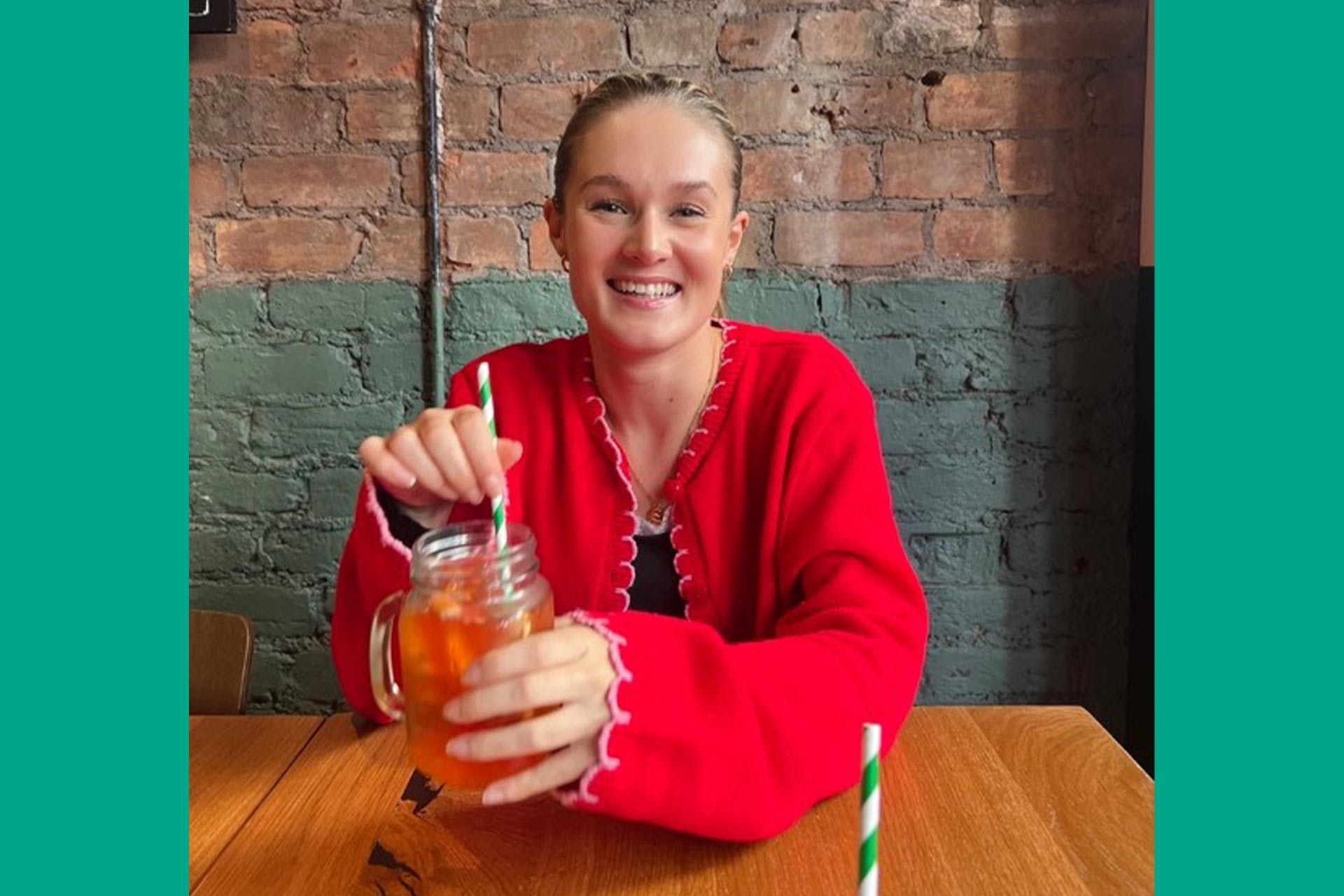 A young woman with blonde hair and red cardigan sitting in a restaurant/bar holding a drink and smiling at the camera.
