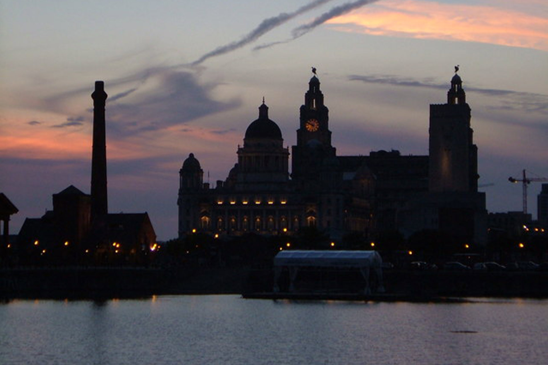 A sun setting on the skyline of Liverpool, which appears dark and shadowy with reflections across the water.