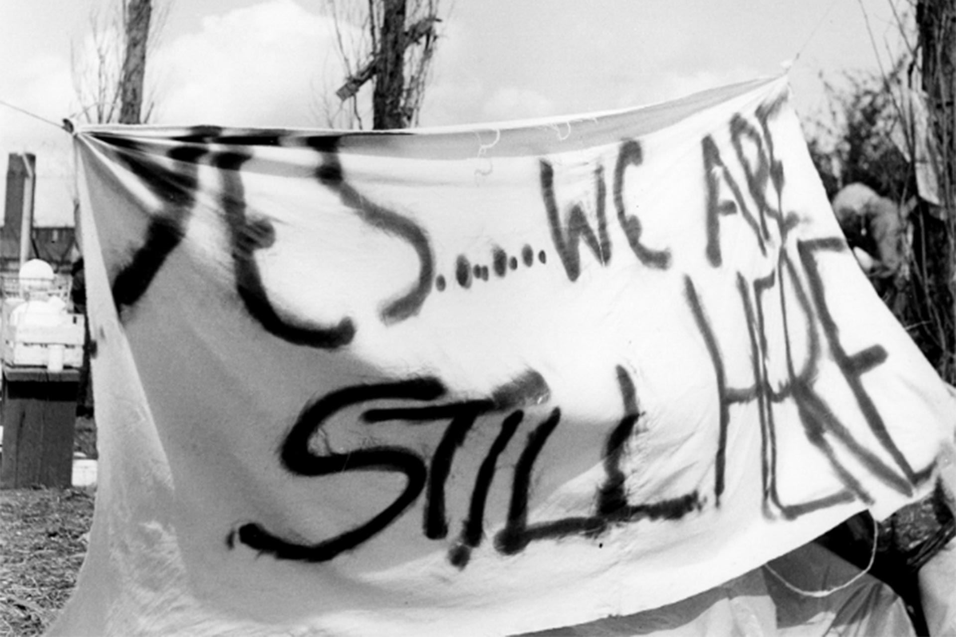 A large white sheet with the message 'Yes... we are still here' handpainted on in large capital letters.