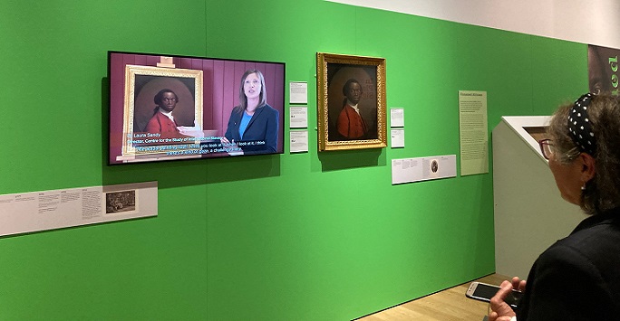 Dr Laura Sandy appearing on a video against a green wall as part of the In Plain Sight exhibition