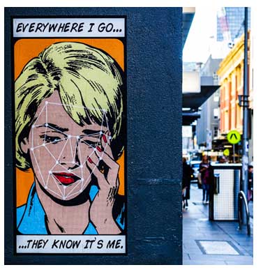 A street scene showing a poster of a comic-book style woman with facial recognition data points over her face, with the words 'Everywhere I go they know it's me'