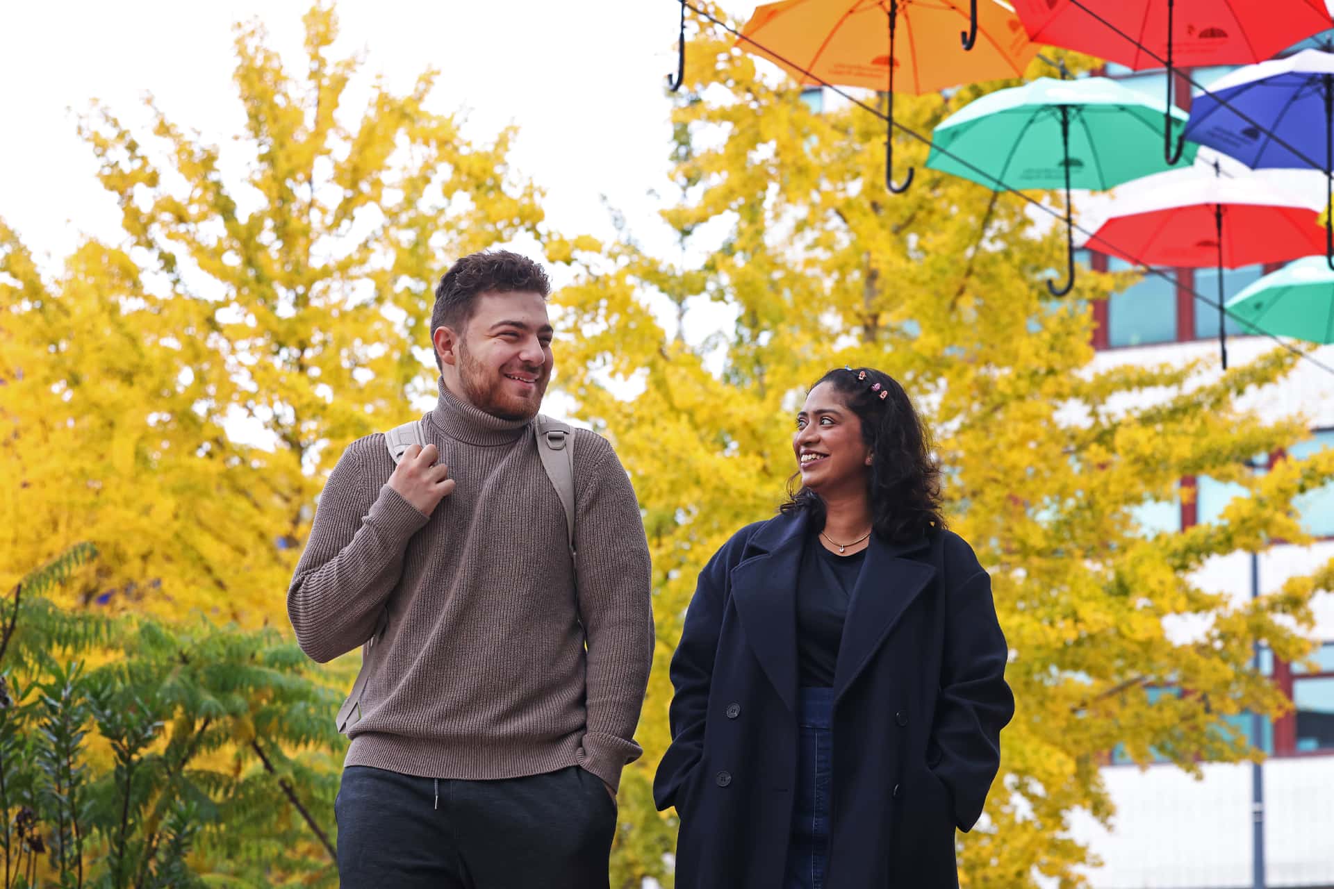 Two people walking along a tree-lined path during autumn.  Above them, colorful umbrellas are suspended in the air.