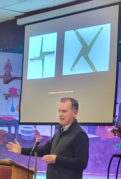 Prof Frank Shovlin standing behind a lectern during his St Brigid's Day talk. Behind him is a PowerPoint presentation showing 2 St Brigid's crosses made from green rushes.