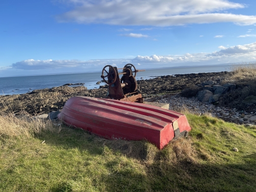 Colour photo of the view from near the Shandwick Stone. In the forground a small red rowing boat is lying upside down on grass, a rusty winch is just behind it. The middle and back of the photo show a rocky shorefront, the sea and blue sky with just a few high clouds.