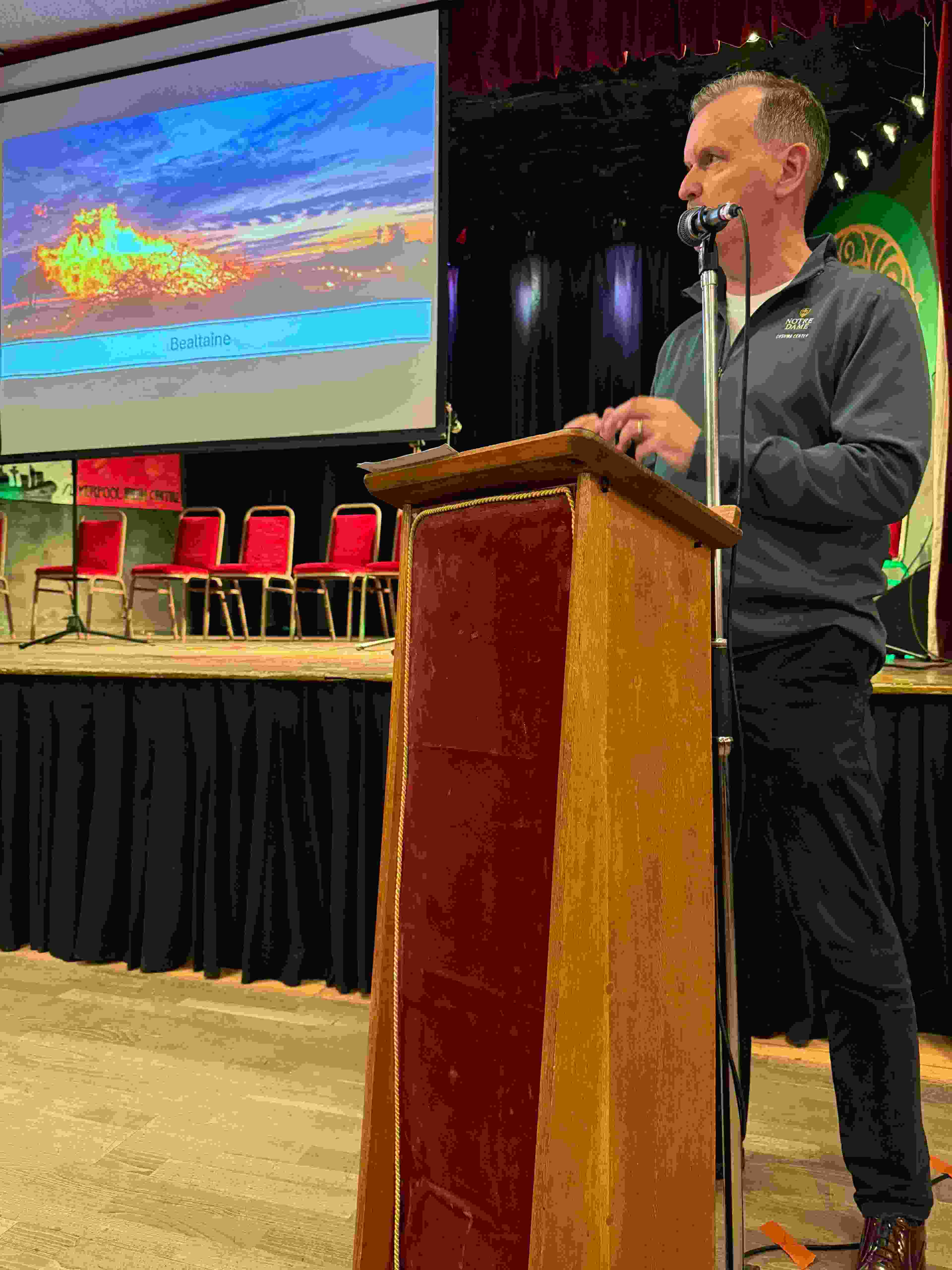 Prof Frank Shovlin standing behind a lectern during his Bealtaine talk.  The slide behind him shows a large bonfire