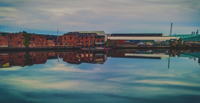 brown industrial style images in a waterfront scene with clouds reflected in the water in front of the buildings 