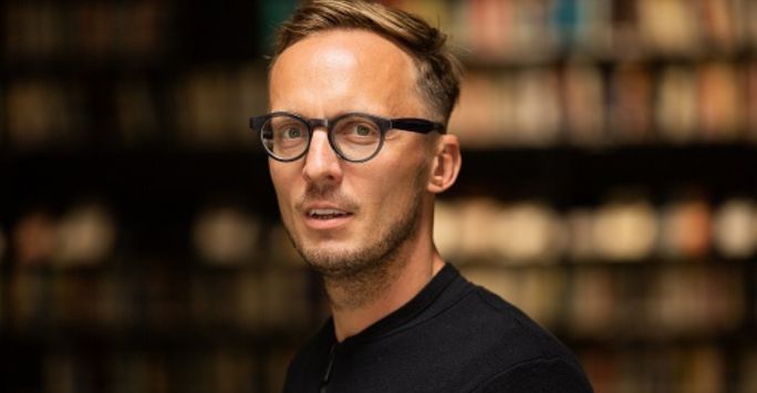 German Academic Michal Hvorecky headshot. A man with black round glasses, a light brown hair and a black top looking straight at the camera. The background is out of focus