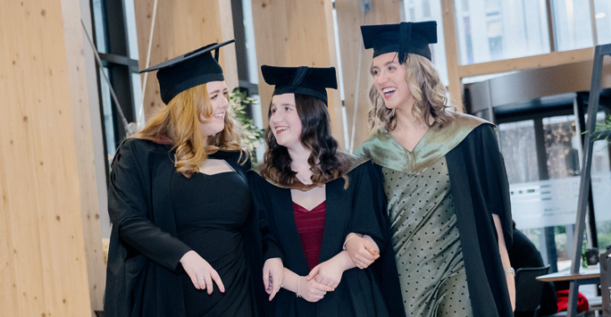 Three law students dressed in graduation gowns and caps with arms linked.