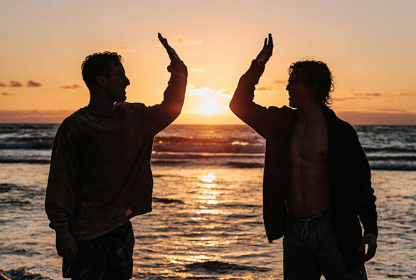 Two men high fiving on a beach in front of a sunset.