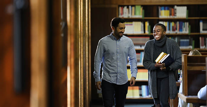 Two students laughing carrying books through the University library.