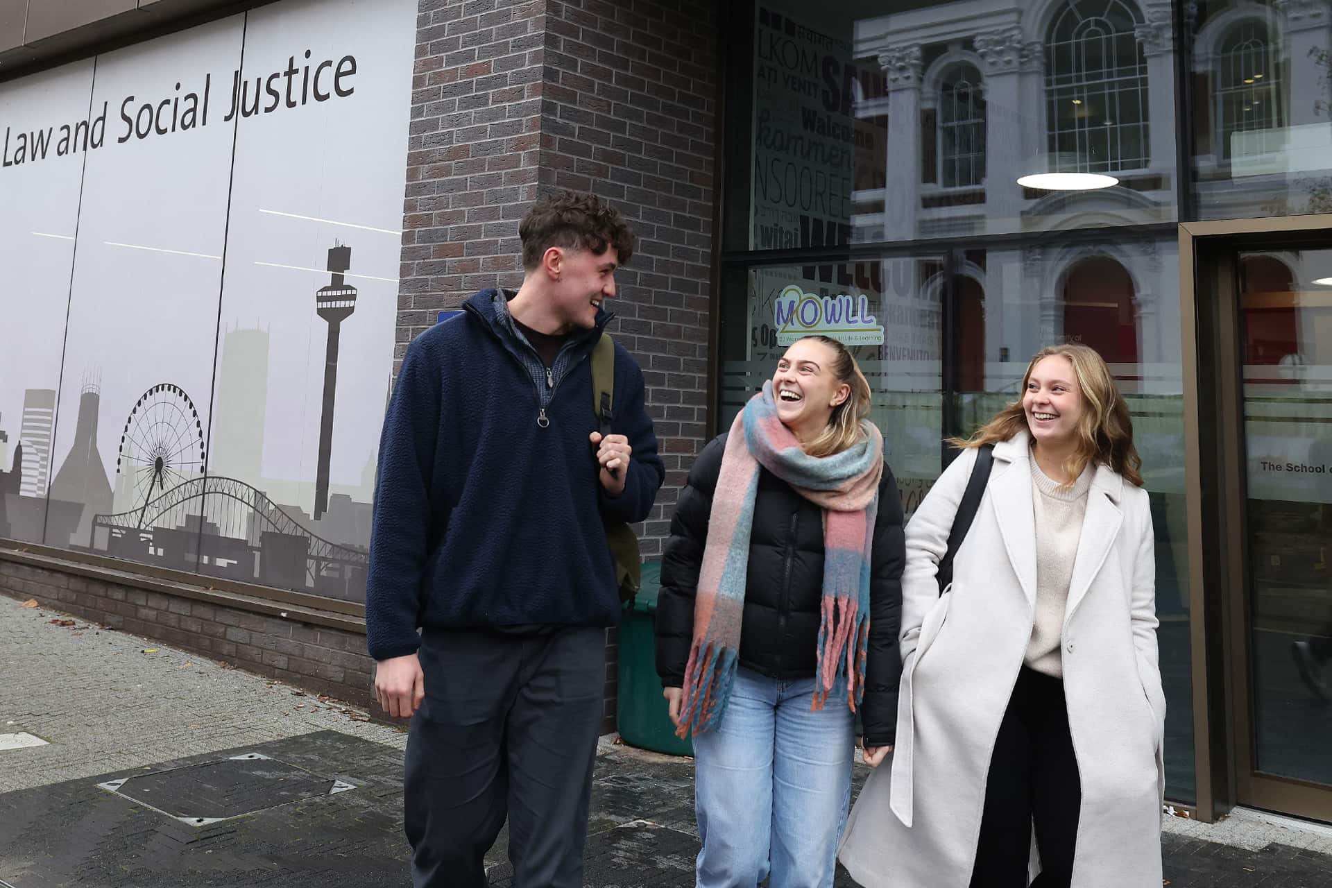 Three smiling students walk together past the School of Law and Social Justice building.