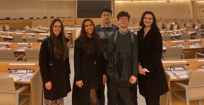 A group of students stood in the Advisory The Human Rights and Alliance of Civilizations Room. The room is filled with brown desks and chairs.