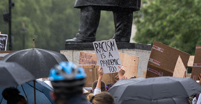 Photo of a placard that reads 'Racism is a Pandemic Too' being held at a Black Lives Matter protest