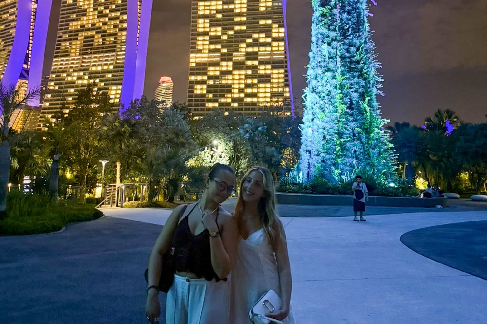 Charlotte Martin and a friend standing in an urban plaza at night, surrounded by tall buildings illuminated with purple lights and a large tree decorated with blue and white lights.