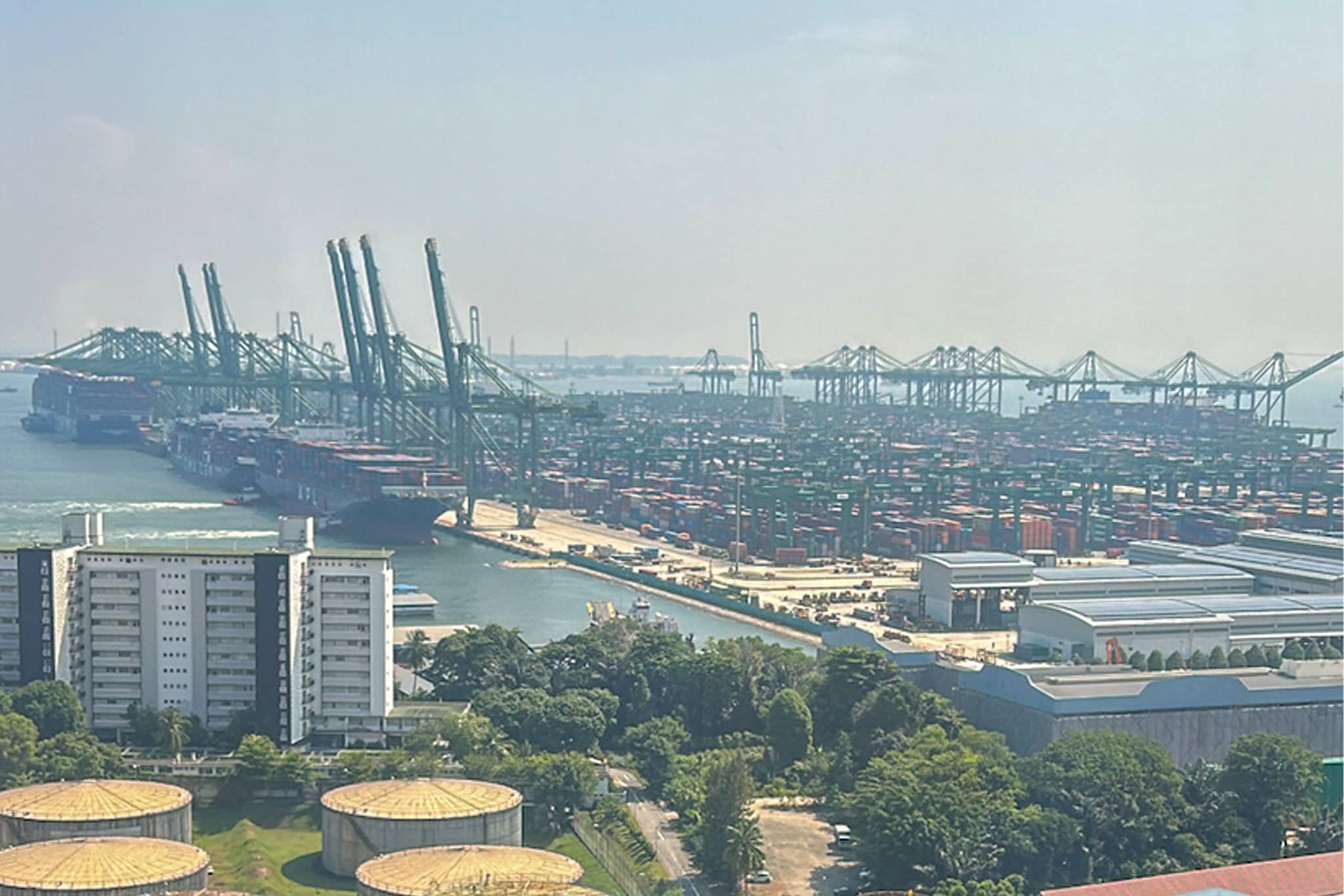 Aerial view of a large port with cargo ships docked, rows of shipping containers, cranes, and circular storage tanks in the foreground, set against a hazy sky over the water.