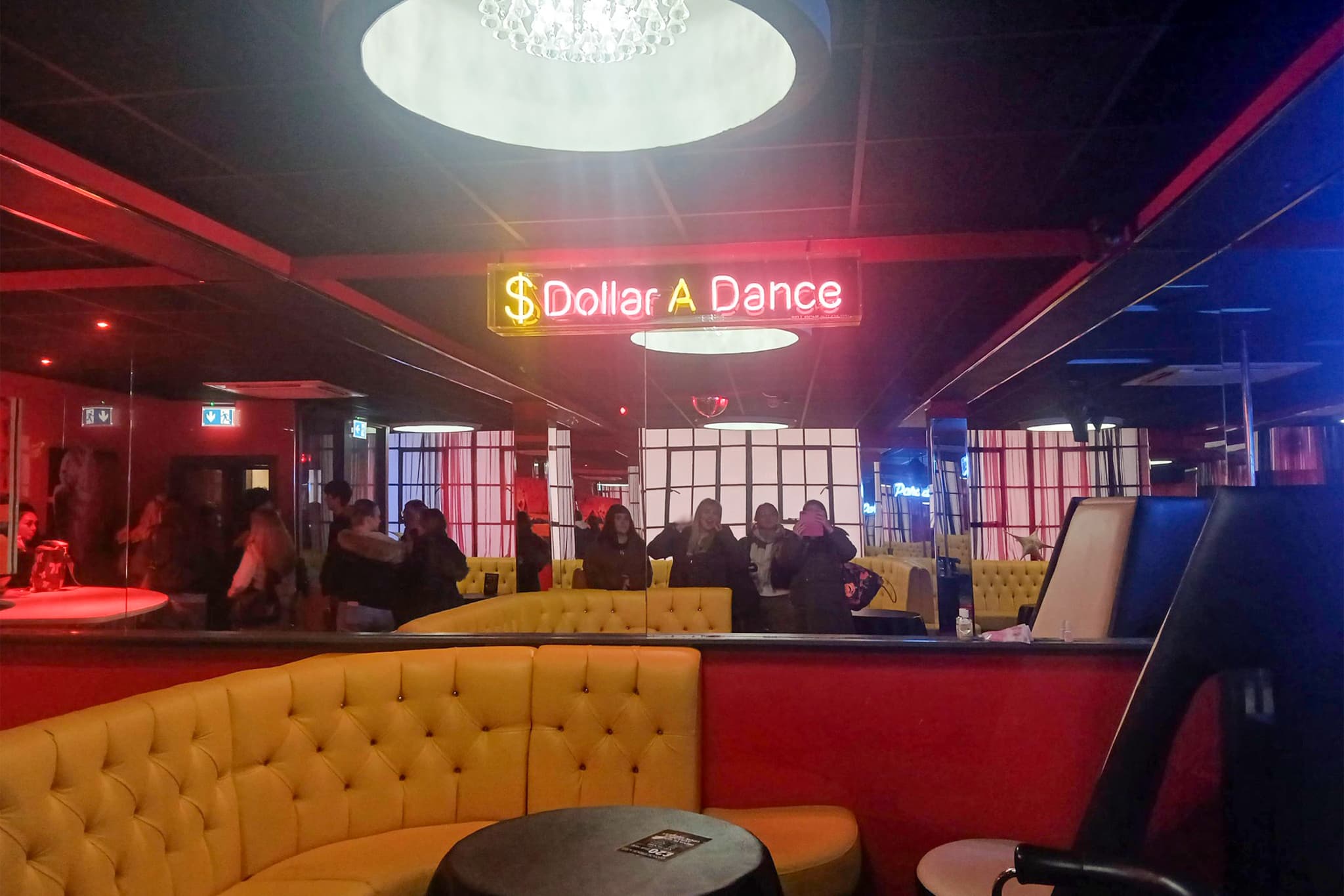 Interior of a sexual entertainment venue with a neon sign reading ‘Dollar A Dance’. A group of students stand near the back of the room, reflected in surrounding mirrors.