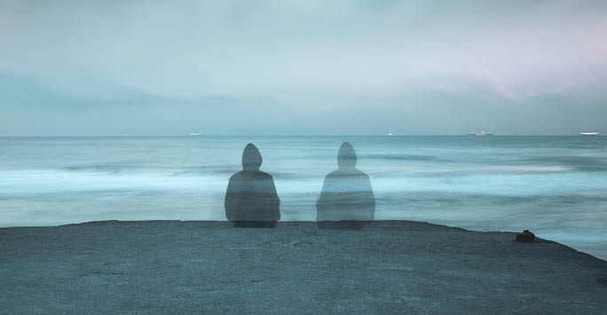 Two people sat in front of the water with one fading away.