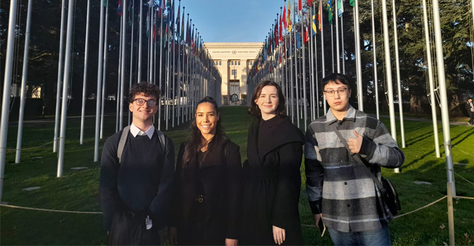 Four students stood outside the United Nations building. There are lots of flag poles with different country flags.