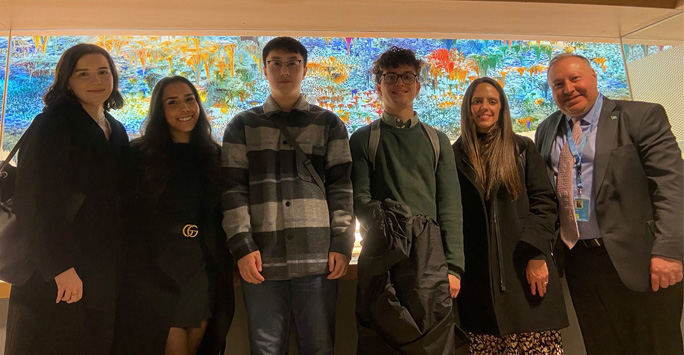 Students and Dr Tzevelekos stand infront a large colourful glass wall.