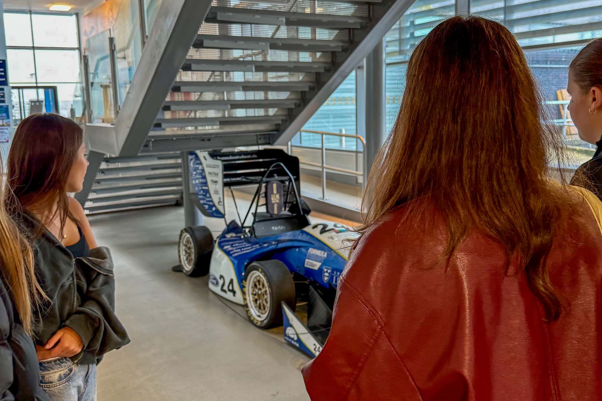 A group of people are observing a blue and white Formula-style race car displayed under a staircase in an indoor setting.