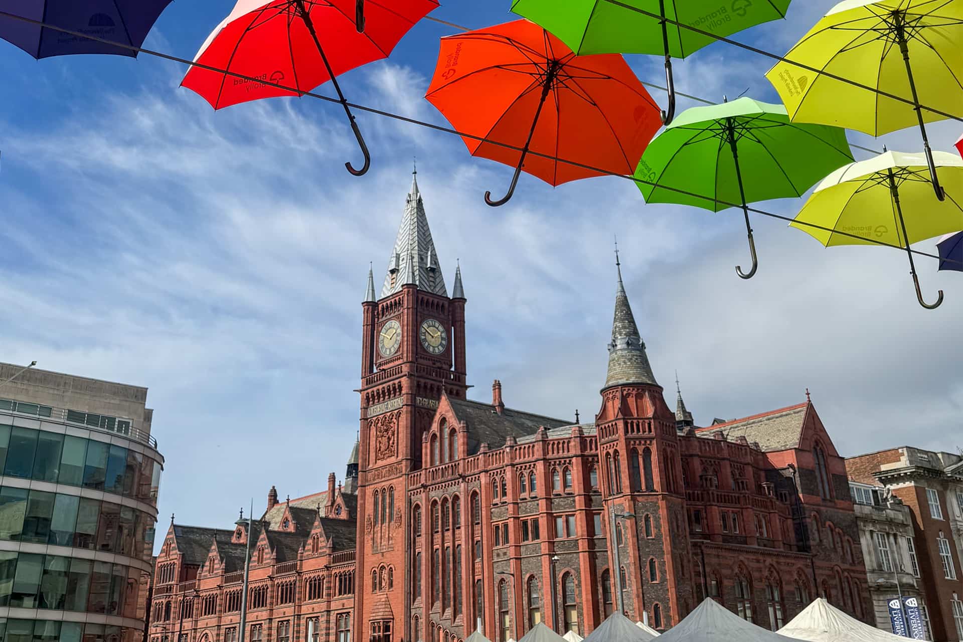 Historic red-brick Gothic-style building with pointed arches, spires. Colorful umbrellas are suspended in the foreground, creating a vibrant canopy.