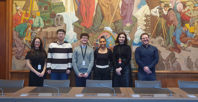 Students stand behind a desk at the World Trade Organisation. Behind them is a large colourful murial.