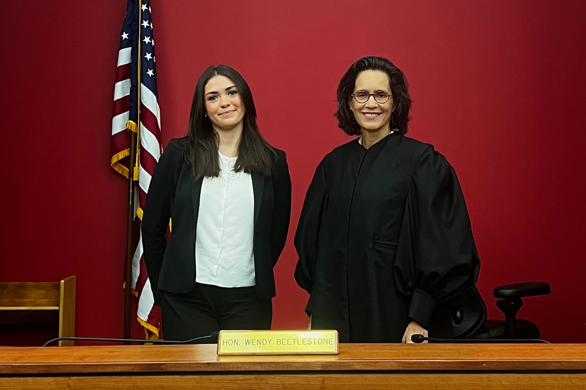 Judge Wendy Beetlestone standing beside intern Sarah in front of a red wall and an American flag, behind a desk with a nameplate reading 'Hon. Wendy Beetlestone.'