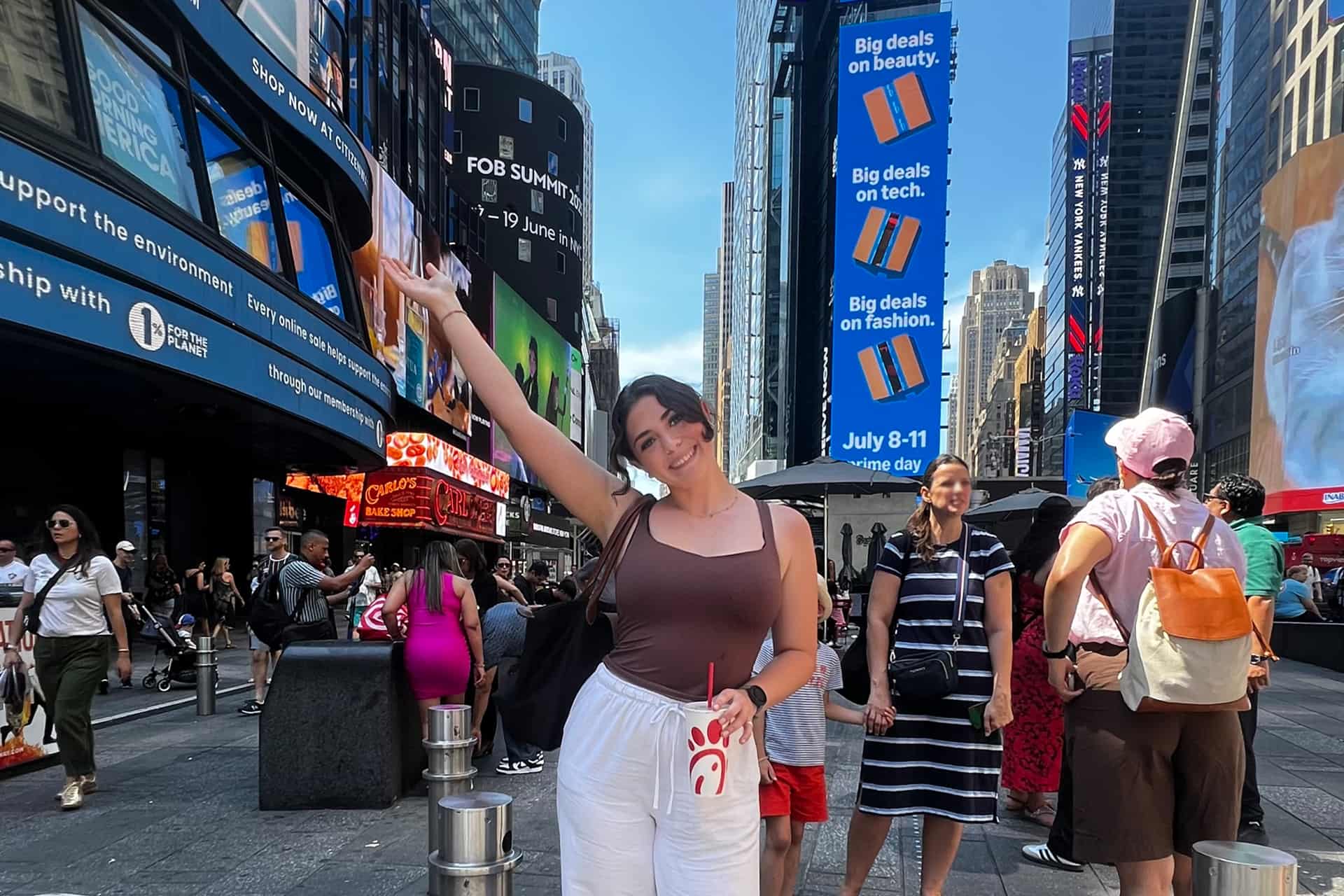 Student Sarah smiles and waves one hand in the air in Times Square, New York City, surrounded by crowds and illuminated digital billboards