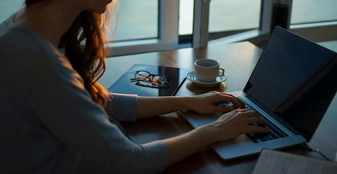 An image of a student sat at a desk with a notepad and an open laptop, writing an essay