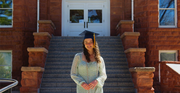 Olivia wearing a graduation cap stood in front of the stairs to a building