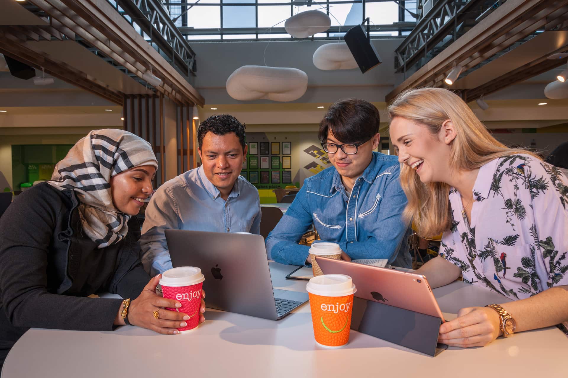 Four people sitting around a table in a modern indoor space, working together on laptops and a tablet with takeaway coffee cups in front of them.