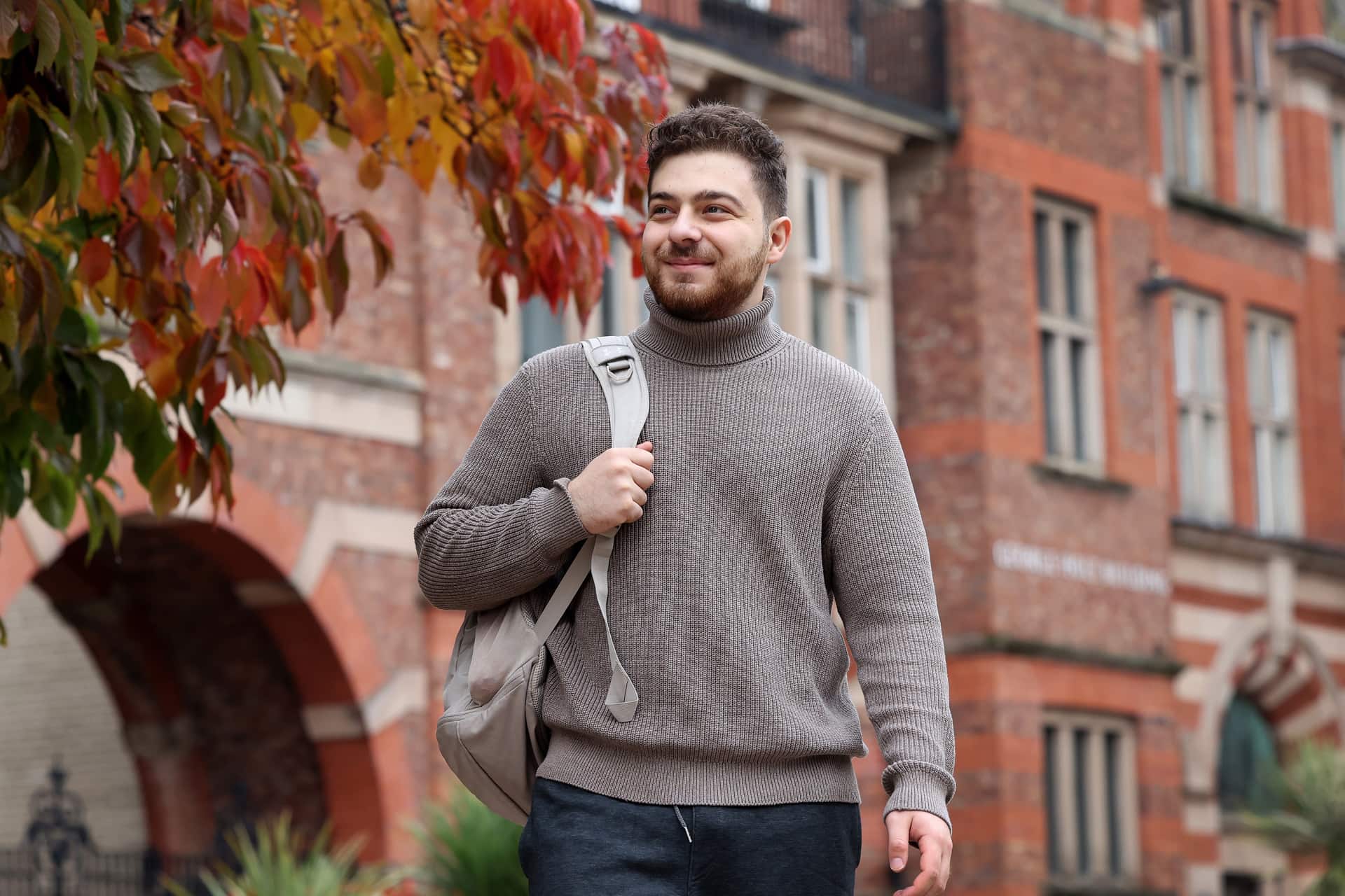 A student walking past a red-brick university building with autumn leaves in the foreground.