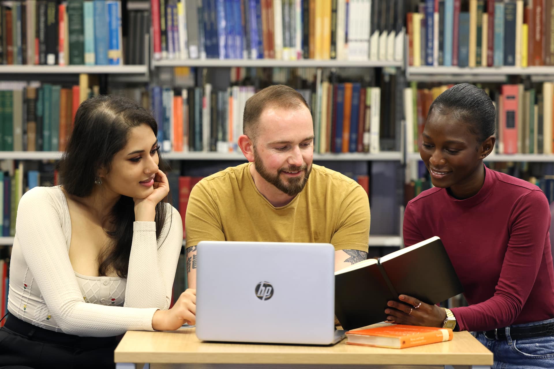Three people sitting at a table in a library, working together with an open laptop and books.