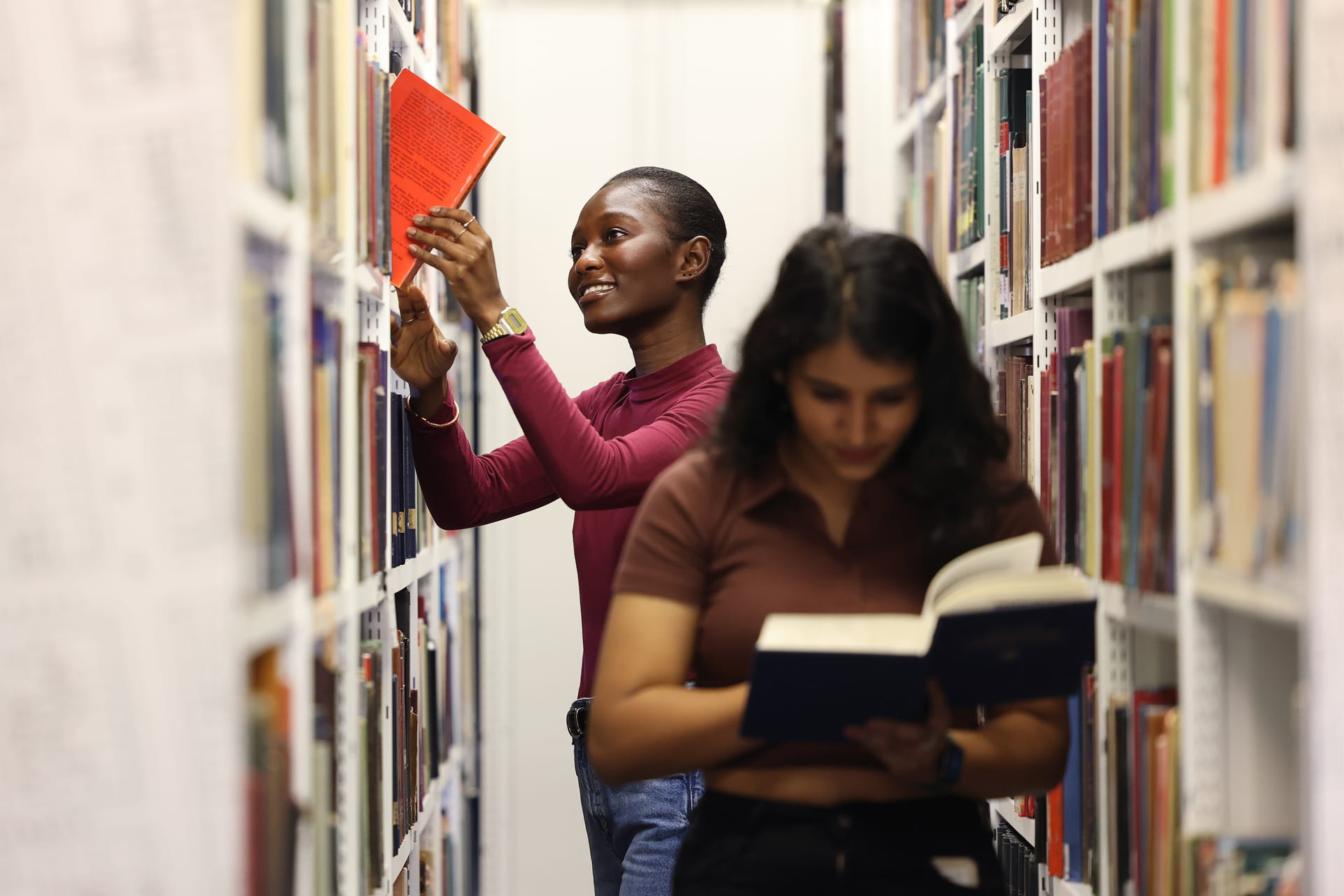 Two students in a library aisle, one reaching for a d book on a high shelf while the other reads a book.