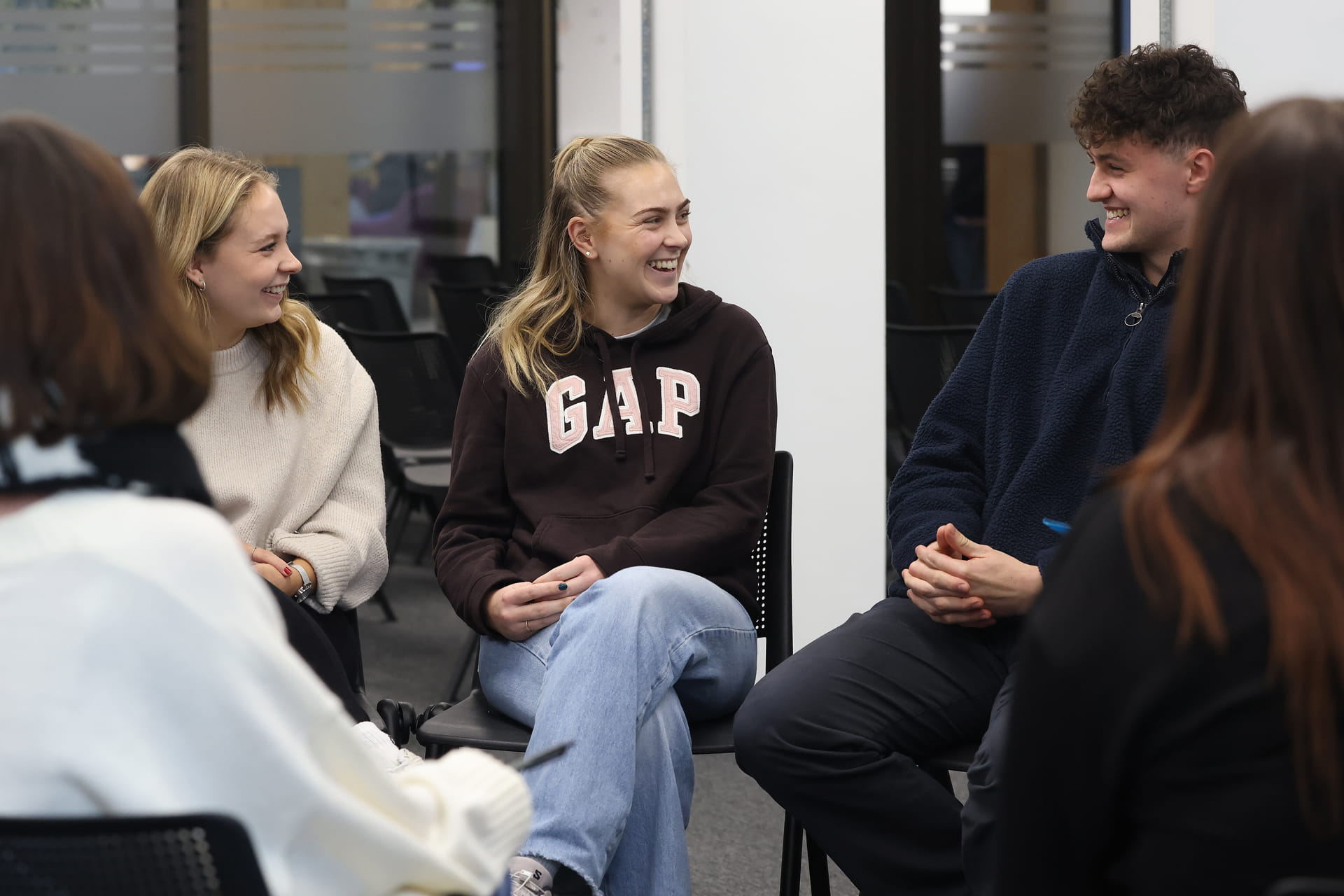 Group of students seated in a circle in an indoor setting, engaged in a discussion.