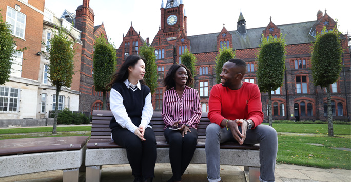 Three students sat on a bench together infront of a university building.