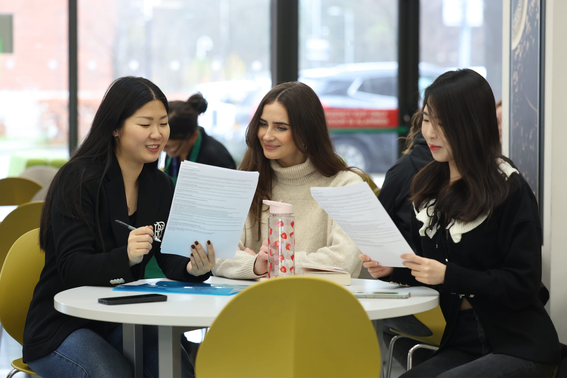 Three people sitting at a round table in a bright indoor space, reviewing printed documents together.
