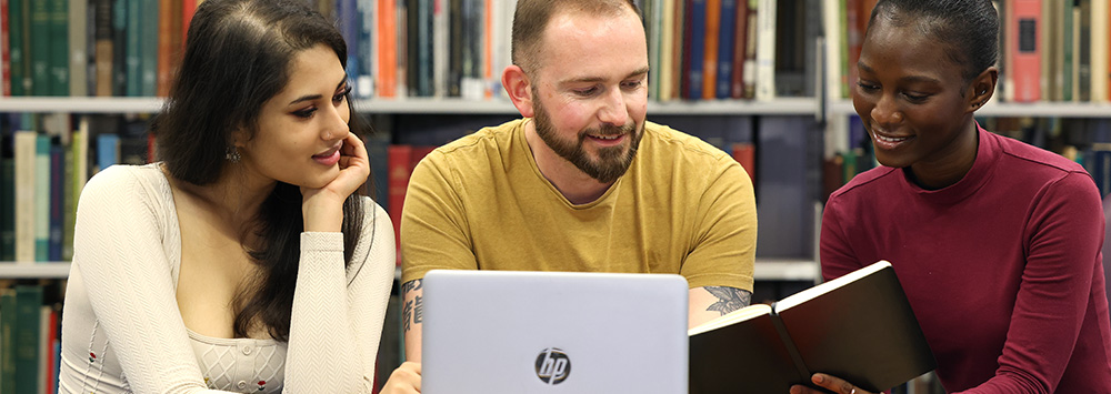 Three students sit in the library looking over a laptop and a book