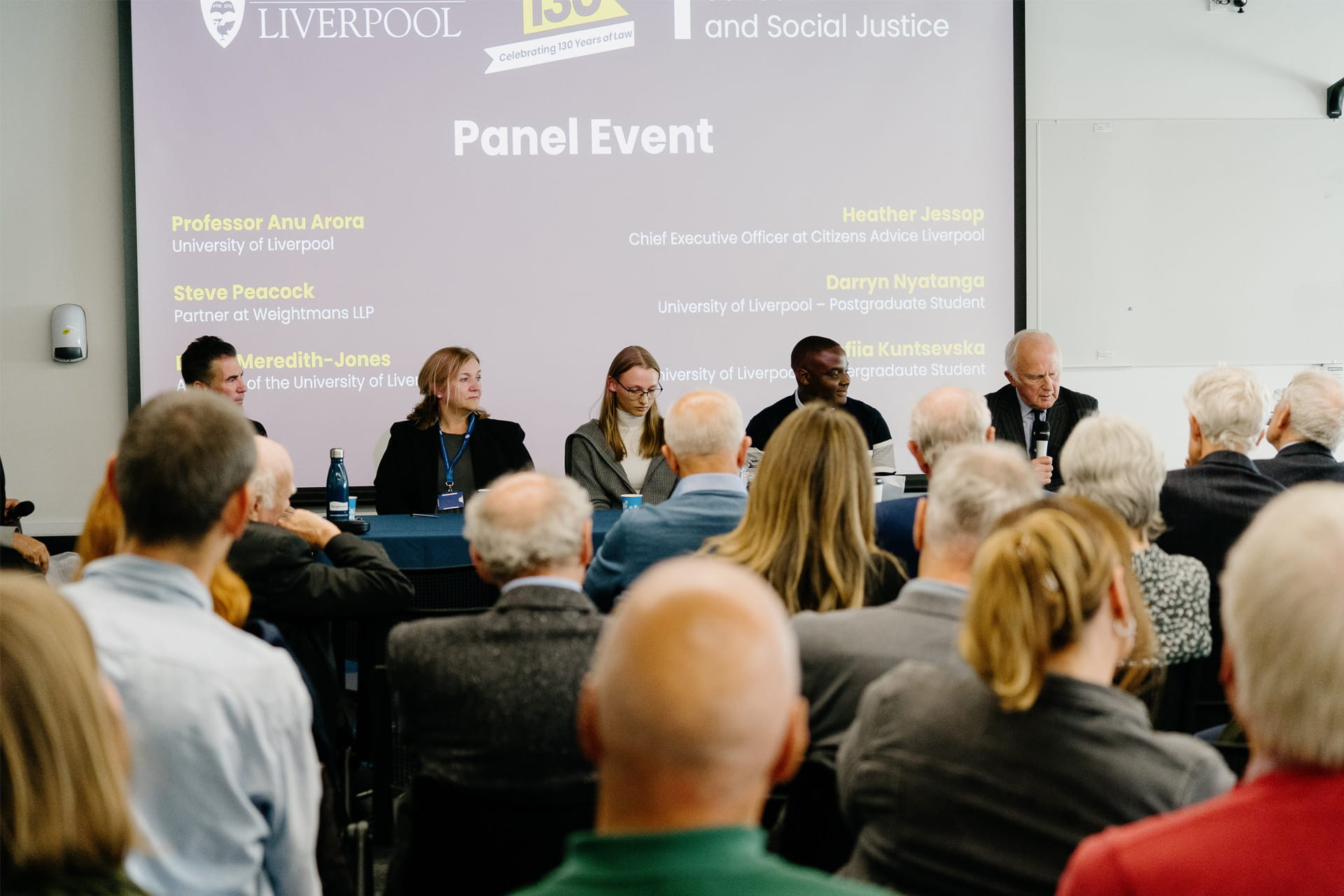 A panel discussion event, with several speakers seated at a long table on a stage in front of a projected screen. An audience sits facing the stage.