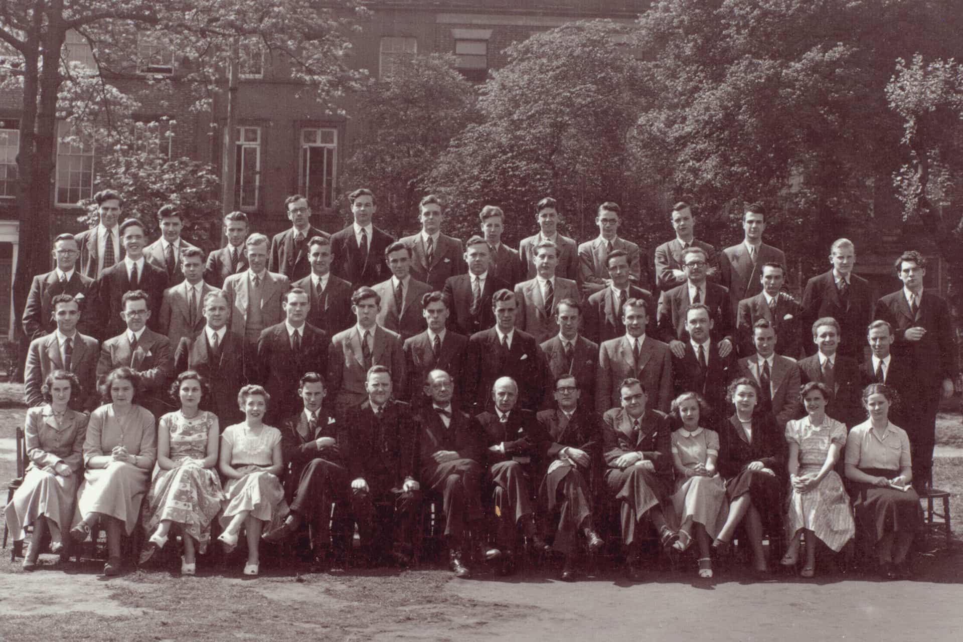 Vintage black‑and‑white class photograph showing a large group of students and staff posed in several rows outdoors, with trees and a building in the background.