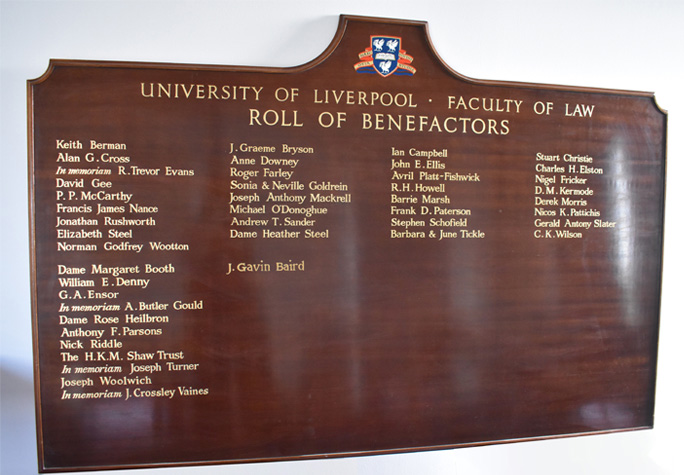 A large wooden plaque mounted on a white wall. The plaque has a list of names written in gold.