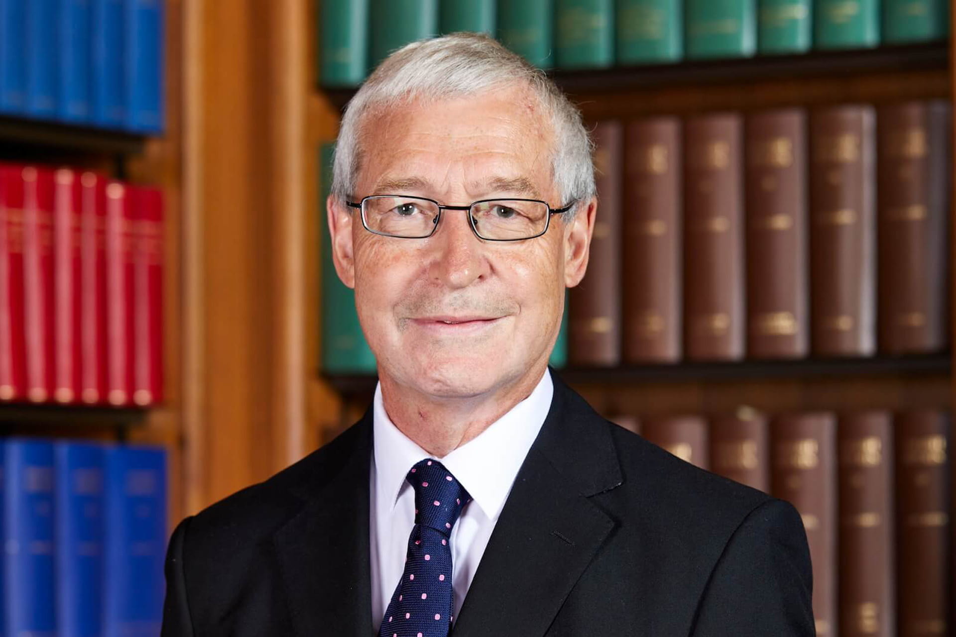 Headshot of Lord Burrows stood in front of a large colourful bookcase.