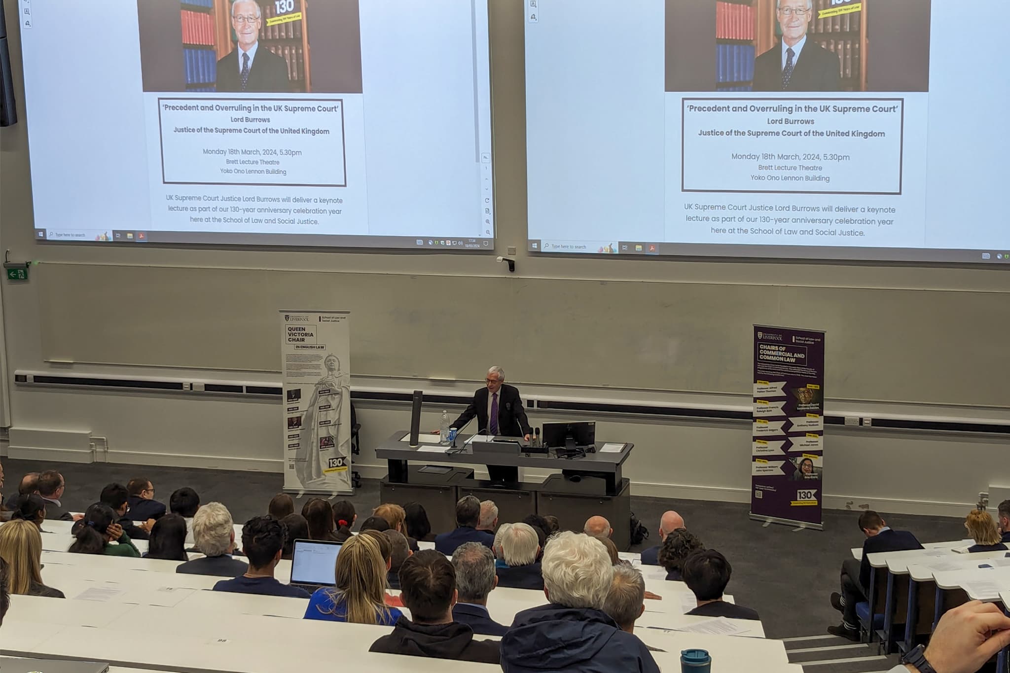 Lord Burrows stands at a lectern in a lecture theatre, addressing an audience seated in tiered rows.