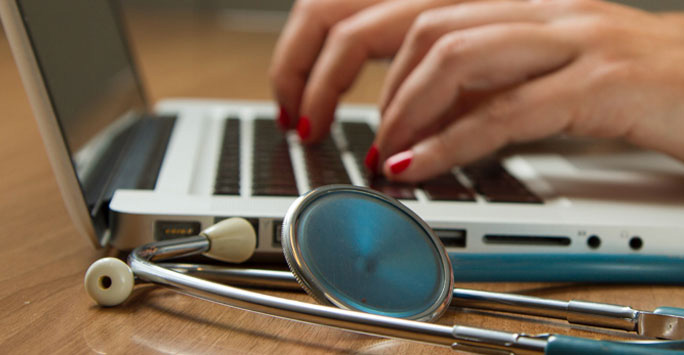 Fingers typing on a laptop next to a stethoscope